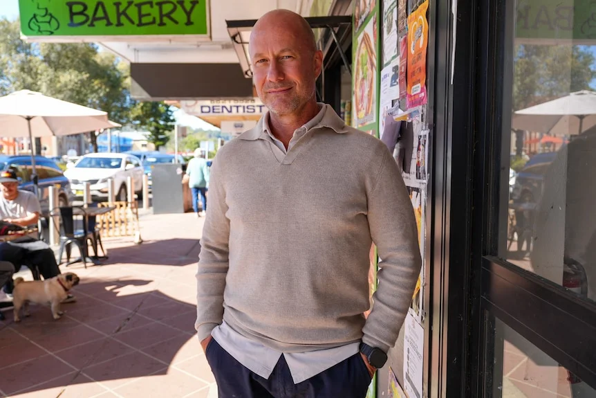 A man iwth a coloured white shirt in a grey knit stands on a street with a cafe in the background.
