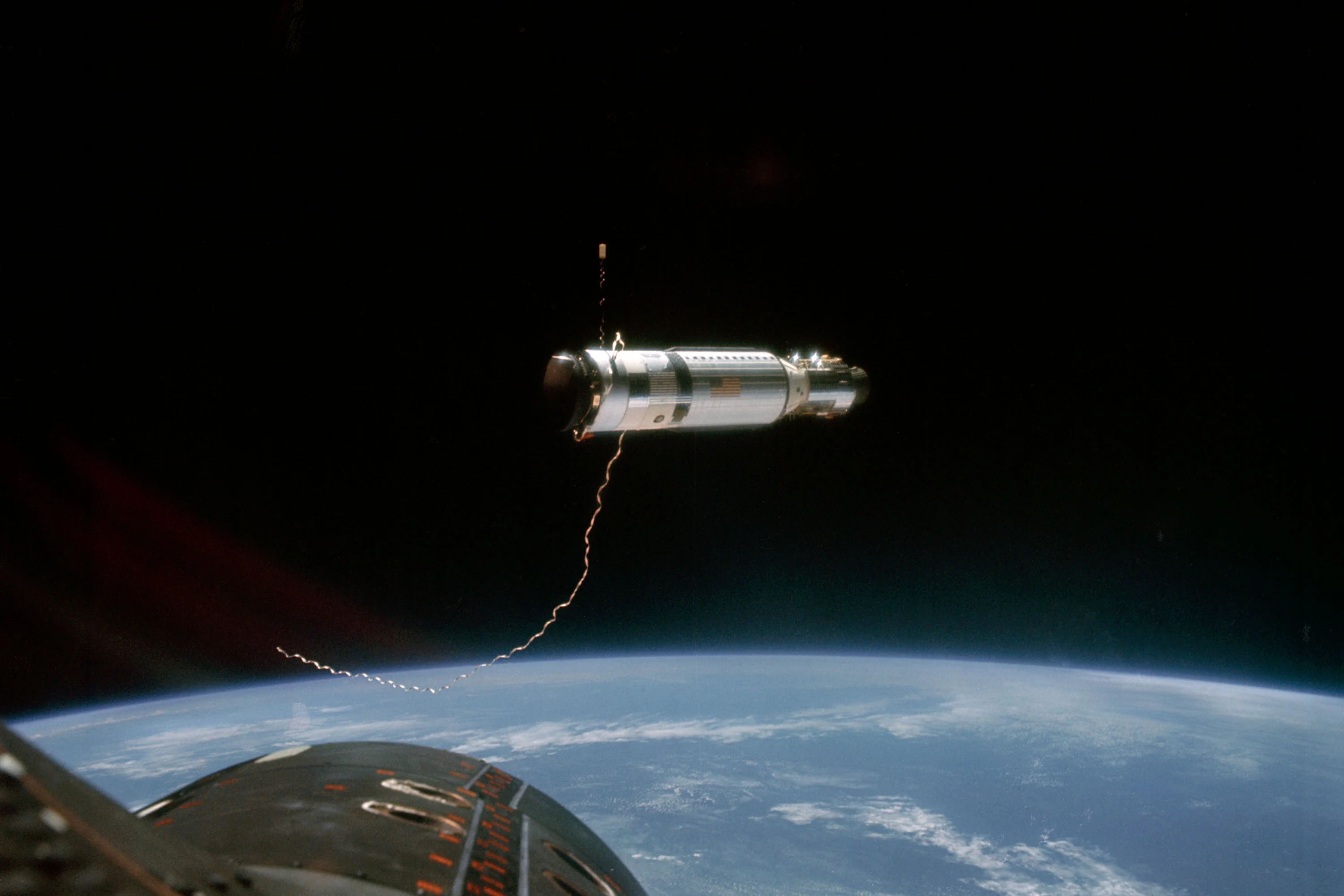 A Gemini spacecraft tethered by an artificial gravity cable to an Agena target vehicle above the Earth’s horizon.